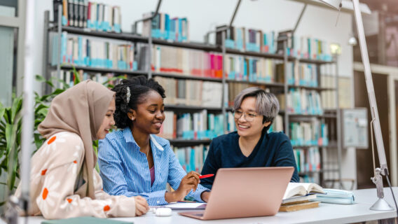 Picture of three people sitting in a library working on a laptop